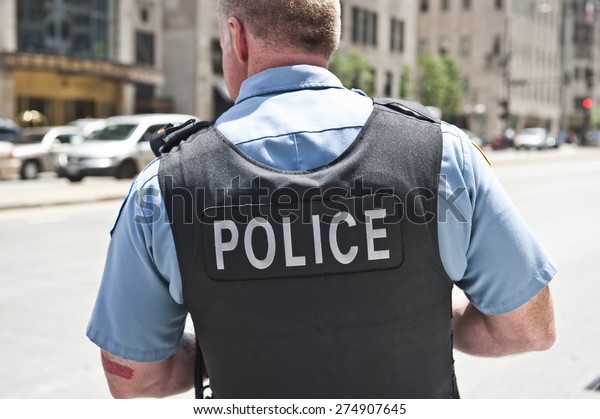 A Chicago\
city policeman standing on the road on a sunny day wearing a bullet\
proof vest mentioning Police on his body. On the background, cars\
and tall buildings are seen.