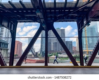 Chicago, IL / USA - 7/19/19: Commuters Walk On Lake Street Underneath El Tracks With Construction Barge Crossing Chicago River In Background During Summer