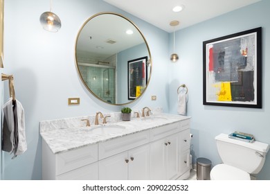 CHICAGO, IL, USA - APRIL 4, 2021: A Light Blue Bathroom With A White Vanity Cabinet, Circular Mirror, Gold Hanging Lights, And Gold Faucets.