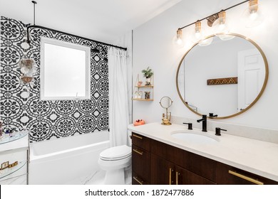 CHICAGO, IL, USA - AUGUST 7, 2019: A Beautifully Renovated Bathroom With A Large Gold Circular Mirror And Light Fixture, Dark Wood Vanity, And A Black And White Pattern Tiled Shower.