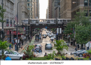 Chicago, IL, USA - Circa 2019: View Down Busy Street In Downtown As Loop Train Passes Overhead Tracks Over Avenue Morning Rush Hour Traffic And Crowds Of People Walking Down Sidewalks To Work