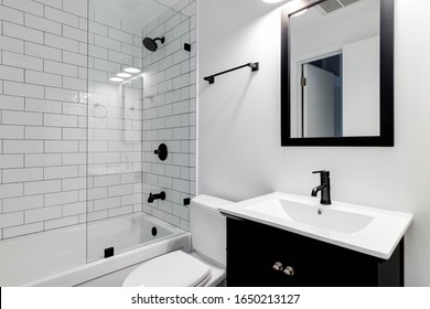 CHICAGO, IL, USA - DECEMBER 27, 2019: A small modern bathroom with a dark vanity, mirror frame, and hardware. White subway tiles line the bathtub and shower with black faucets.