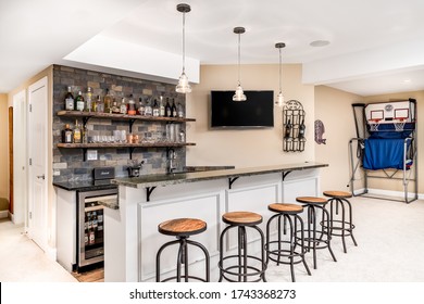 CHICAGO, IL, USA - FEBRUARY 24, 2020: A Small Basement Bar /man Cave With Liquor On Shelves And The Bar Is Surrounded By Wood Bar Stools. A Basketball Game Is Setup In The Background.