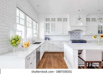 CHICAGO, IL, USA - JULY 21, 2020: A luxury white kitchen with bar stools sitting at a large island, glass lights hanging from the ceiling, and a beautiful tiled backsplash.