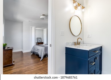 CHICAGO, IL, USA - JUNE 6, 2020: A Bright White Bathroom With A Blue Vanity, Gold Light Fixture, And Gold Circular Mirror Look Looking Towards A Large Bedroom With Hardwood Floors. 