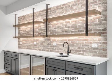 CHICAGO, IL, USA - OCTOBER 15, 2020: A Basement Wet Bar With A White Granite Countertop, Grey Cabinet, Lighting Under The Shelves Mounted To Exposed Brick, And Built-in Refrigerators.