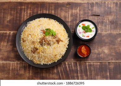 Chicken biryani , kerala wedding chicken dhum biriyani made using jeera rice  with raitha and lemon pickle as side dish arranged in a black table ware with wooden background,top view.