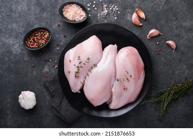 Chicken breast. Raw chicken breast fillets on black ceramic plate on wooden cutting board with herbs and spices on black background. Top view with copy space.
