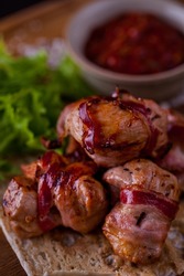 Chicken Fillet Shashlik, Wrapped In Bacon With Homemade Adjika In A Beige Saucepan. The Kebab Lies On A Sheet Of Pita Bread, Next To It Are Pickled Onions And Lettuce. Food And Sauce Lie On A Wooden B