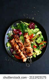 Chicken kebab skewers with fresh vegetable salad on a black plate over dark slate, stone or concrete background. Top view with copy space.