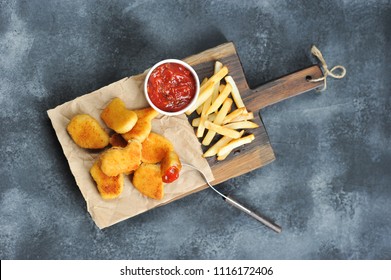 Chicken nuggets and French fries on a wooden board. Next cup with ketchup. One of the nuggets is pinned on the fork. View from above. Close-up. Gray background.