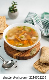 
chicken soup broth with vegetables in a white bowl, next to a spoon, bread, towel, spices