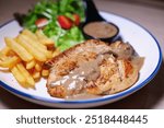 Chicken steak with side dishes of French fries, salad and pepper sauce on a white plate, photographed with studio lights.