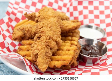 Chicken Tenders Breaded And Cooked To Perfection Sitting On Top Of A Pile Of Waffle French Fries Along With Dipping Sauces.