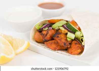 Chicken Tikka Wrap - Tandoori chicken tikka with salad wrapped in a flatbread, served with chili sauce, yogurt and mint raita and lemon wedges on a white background.