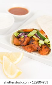 Chicken Tikka Wrap - Tandoori chicken tikka with salad wrapped in a flatbread, served with chili sauce, yogurt and mint raita and lemon wedges on a white background.