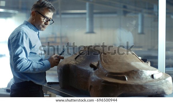 Chief Automotive Designer with Rake Sculpts\
Futuristic  Car Model from Plasticine Clay. He Works in a Special\
Studio Located In a Large Car\
Factory.