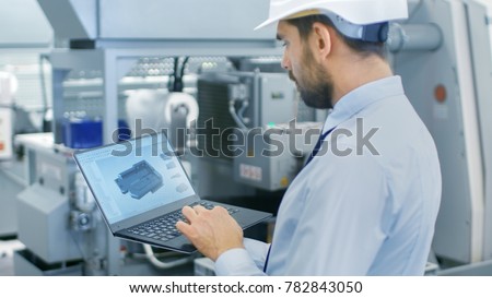 Chief Engineer in the Hard Hat Holds Laptop with 3D Component Model on it's Screen. In the Background Modern Factory Equipment.