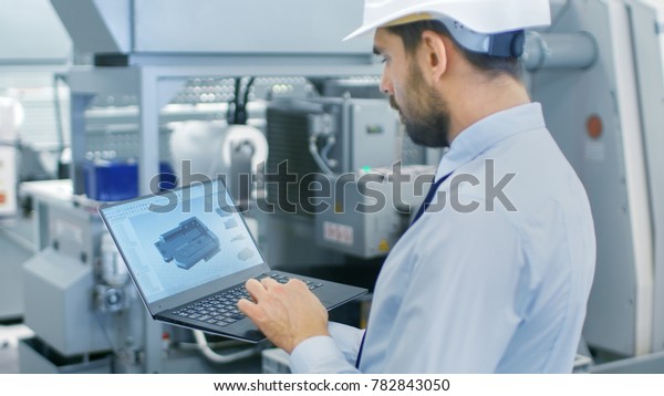 Chief Engineer in the Hard Hat Holds Laptop\
with 3D Component Model on it\'s Screen. In the Background Modern\
Factory Equipment.