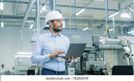 Chief Engineer in the Hard Hat Walks Through Light Modern Factory While Holding Laptop. Successful, Handsome Man in Modern Industrial Environment.