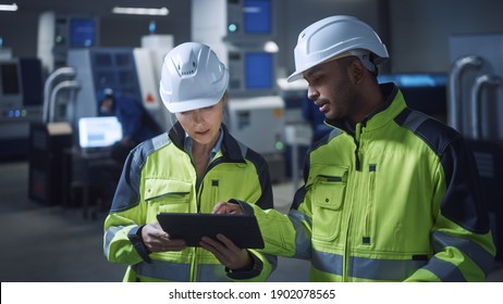 Chief Engineer and Project Manager Wearing Safety Vests and Hard Hats, Use Digital Tablet Computer in Modern Factory, Talking, Optimizing Production Line. Industrial Facility with CNC Machinery