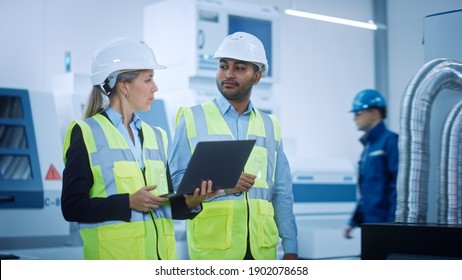 Chief Engineer And Project Manager Wearing Safety Vests And Hard Hats Walk Through Modern Factory, Talking, Optimizing Production Line. Industrial Facility: Professionals Working On Machinery