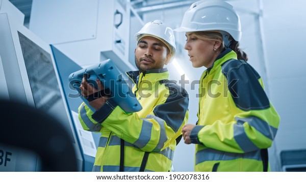 Chief Engineer and Project Manager Wearing\
Safety Vests and Hard Hats, Use Digital Tablet Controller in Modern\
Factory, Talking, Programming Machine For Productivity. Low Angle\
Portraits