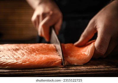 Chief hands cut salmon fillet with knife on wooden table at kitchen. Man cooking red omega fish with lemons for healthy nutrition diet