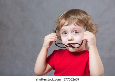 A child of 3 years old  with long hair tries to put on  eye glasses. Gray background  Free space for a text.