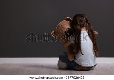 Child abuse. Upset little girl with teddy bear sitting on floor near gray wall indoors, back view. Space for text
