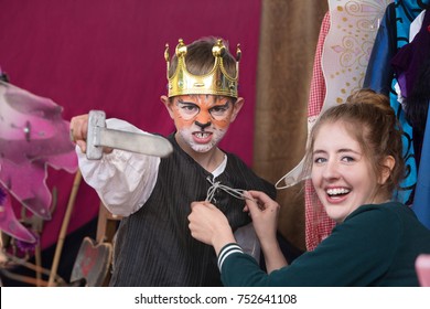 Child Actor Dressed As King With Crown Brandishes Sword As Assistant Helps Him 