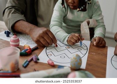 Child and adult engaging in Easter egg drawing activity using crayons and paper on wooden table. Hands of both participants carefully working on intricate designs - Powered by Shutterstock