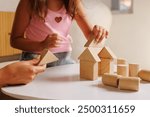 A child and an adult are playing with wooden building blocks on a white table. The child is placing a triangular block on top of a square block, while several other blocks are scattered around