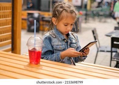 A Child With Afro-pigtails In A Denim Jacket Is Playing Games On A Smartphone. The Kid Is Sitting At A Table In A Patio In A Cafe And Looks Intently At The Phone. A Girl Drinks Lemonade. Summer Day