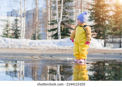 A Child Aged 2-3 Years Old Dressed In A Yellow Rubber Jumpsuit Stands In A Puddle In A City Park In The Spring