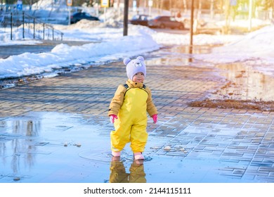 A Child Aged 2-3 Years Old Dressed In A Yellow Rubber Jumpsuit Runs Through Puddles In A City Park In Early Spring