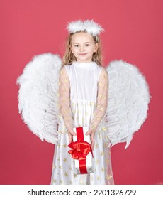 Child angel little girl with present gift, studio portrait. Little angel with white wings holds gift. Valentine gift card. Cute angel child girl with angels wings, isolated on red. Valentines day.