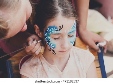 Child animator, artist's hand draws face painting to little girl. Child with funny face painting. Painter makes blue butterfly at girl's face. Children holiday, event, birthday party, entertainment. - Powered by Shutterstock