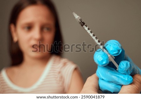 A child appears apprehensive while observing a healthcare worker holding a syringe in a gloved hand. The setting suggests a medical facility focused on vaccination.