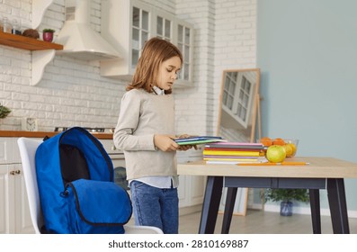 Child arranges a stack of colorful school books beside a blue backpack on the kitchen table, preparing notebooks and supplies for school in a bright home. She packs books for school now.