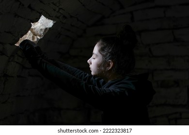 A Child In A Basement Looking In A Small Window. A Beam Of Light Falls On Her Face. Portrait Of The Little Girl In The Dungeon With Dark Background.