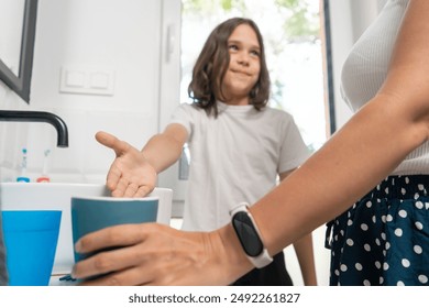 The child in the bathroom points to a rinsing cup, suggesting to use it. The mother stands nearby, observing to ensure the morning routines of brushing teeth and washing the face are done correctly. - Powered by Shutterstock