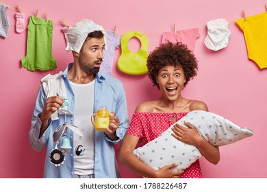 Child Birth, Parenting, Fatherhood And Motherhood Concept. Young Busy Parents With Infant, Busy Multitasking Father Feeds Baby, Going To Change Nappy, Holds Mobile, Stares At Crying Newborn.