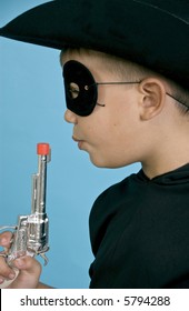A child in a black cowboy hat and mask, blowing on the end of a toy gun, as if just discharged.