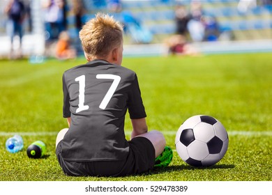 Child In Black Sportswear With Soccer Ball. Boy Sitting On Football Pitch. School Football Match For Children