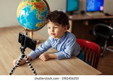 A Child Blogger Shoots A Video On A Smartphone On A Tripod. The Boy Poses On A Smartphone Camera, Shoots A Vlog, Next To The Globe. Funny Cute Kid