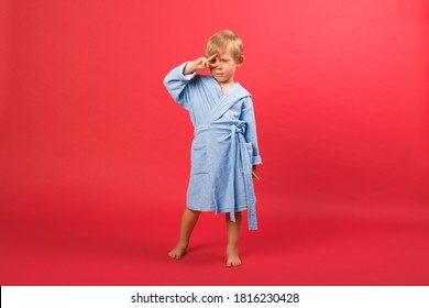 Child Blond Boy In A Blue Bathrobe On A Red Bright Background Shows Sign Peace, Victory Disco Near The Eyes. Studio Shot Of Kid Dancing To Advertise A Bathhouse, Sauna, Spa, Pool And Swimming School.