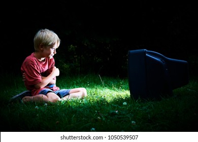 Child Blond Boy Watching TV Horror / Thriller. Outside In The Garden At Night. Concept Of Inappropriate Movies For Children
