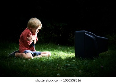 Child Blond Boy Watching TV Horror / Thriller. Outside In The Garden Sitting On Grass Lawn At Night. Concept Of Inappropriate Movies For Children
