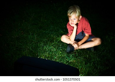 Child Blond Boy Watching TV Horror / Thriller. Outside In The Garden Sitting On Grass Lawn At Night. Concept Of Inappropriate Movies For Children